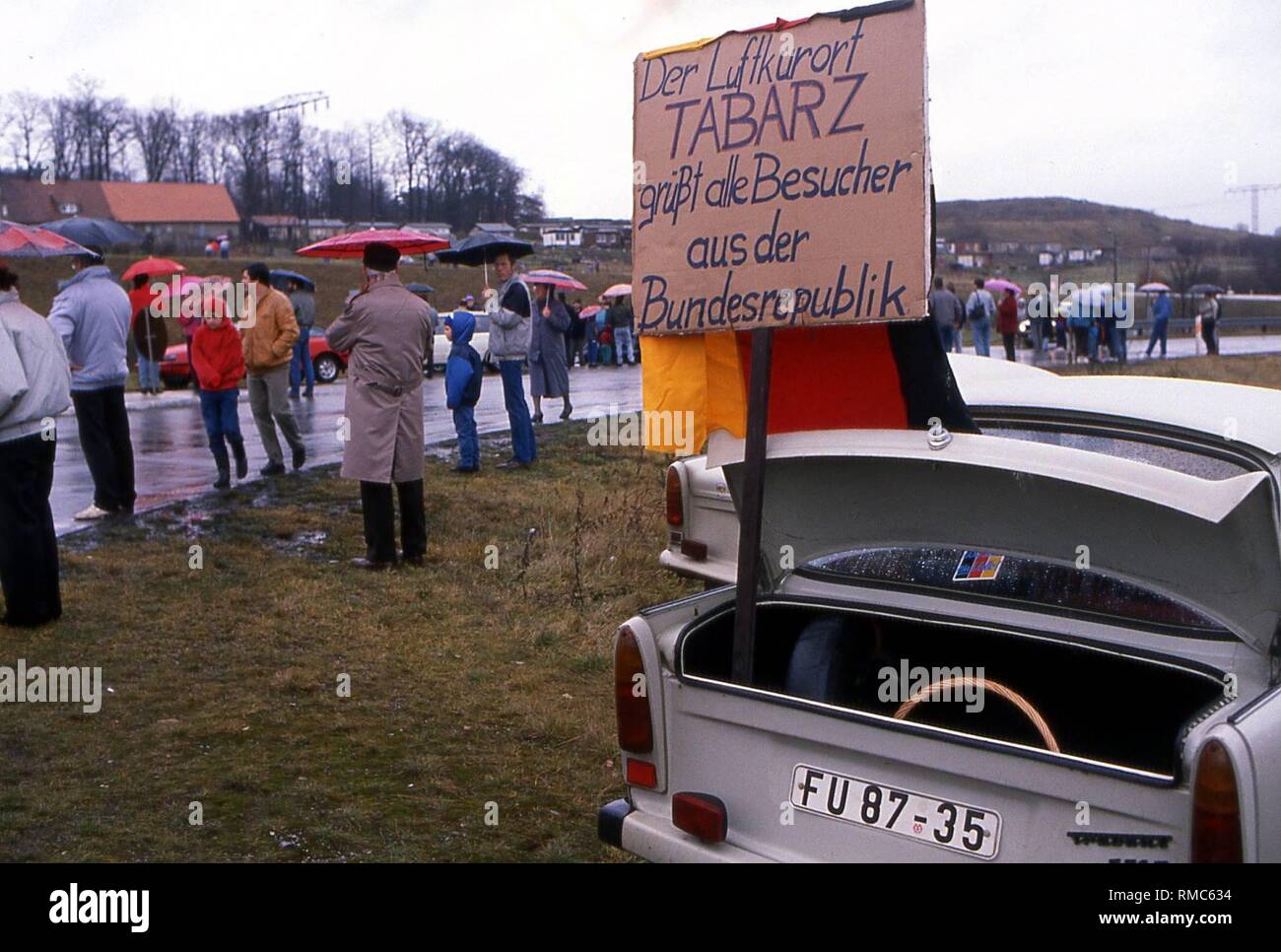 A sign is in the trunk of a Trabant on the roadside in Tabarz, which says 'The climatic health-resort Tabarz greets visitors from the Federal Republic'. In the background are people with umbrellas. Stock Photo