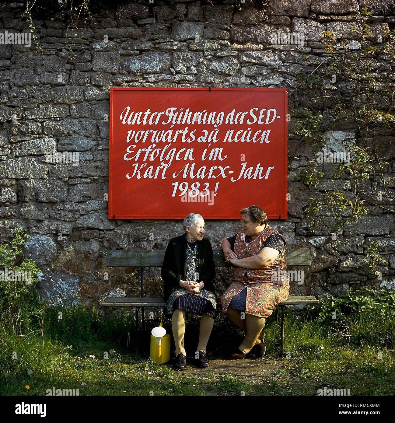 Two old women are sitting on a bench in a village in Thueringen. On the wall beyond them a propaganda- sign to the Karl- Marx- Year. Stock Photo