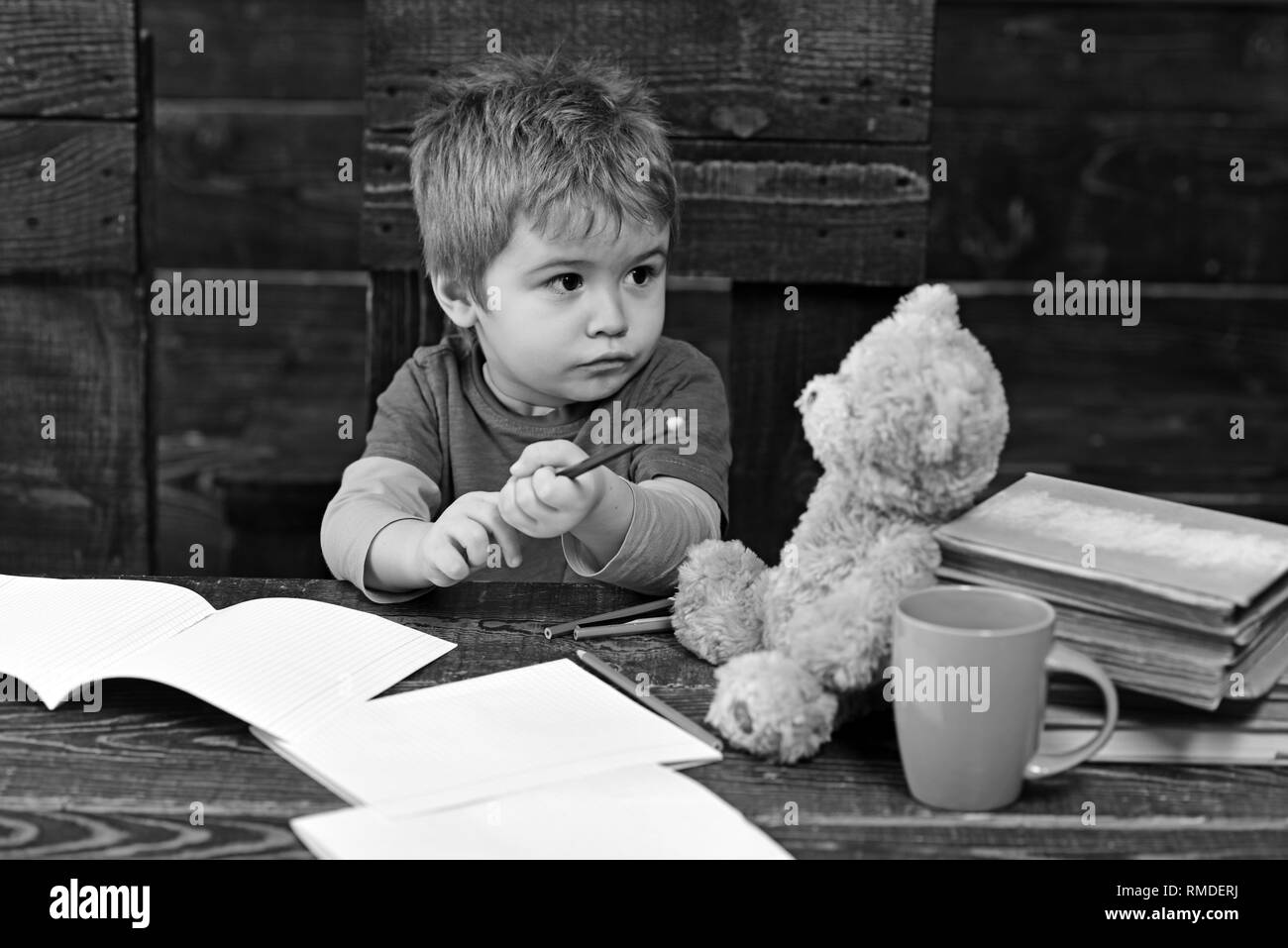 Busy kid at school. Small boy sitting at wooden table with copybooks. Blond child looking at his teddy bear Stock Photo