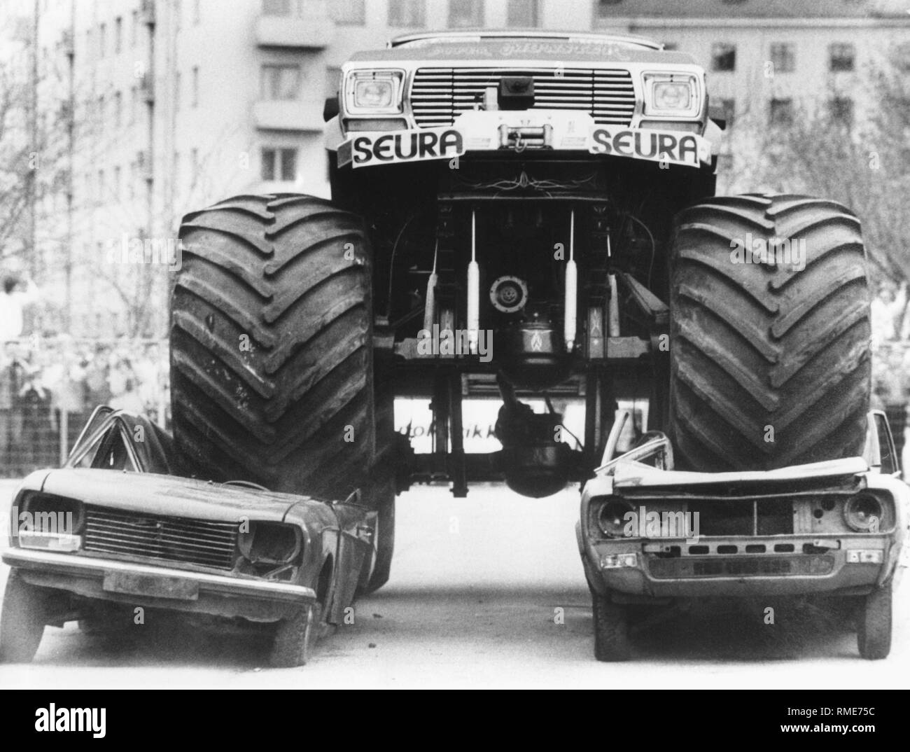 A monster truck in Helsinki drives over two car bodies. On the left, a Peugeot 504 Stock Photo