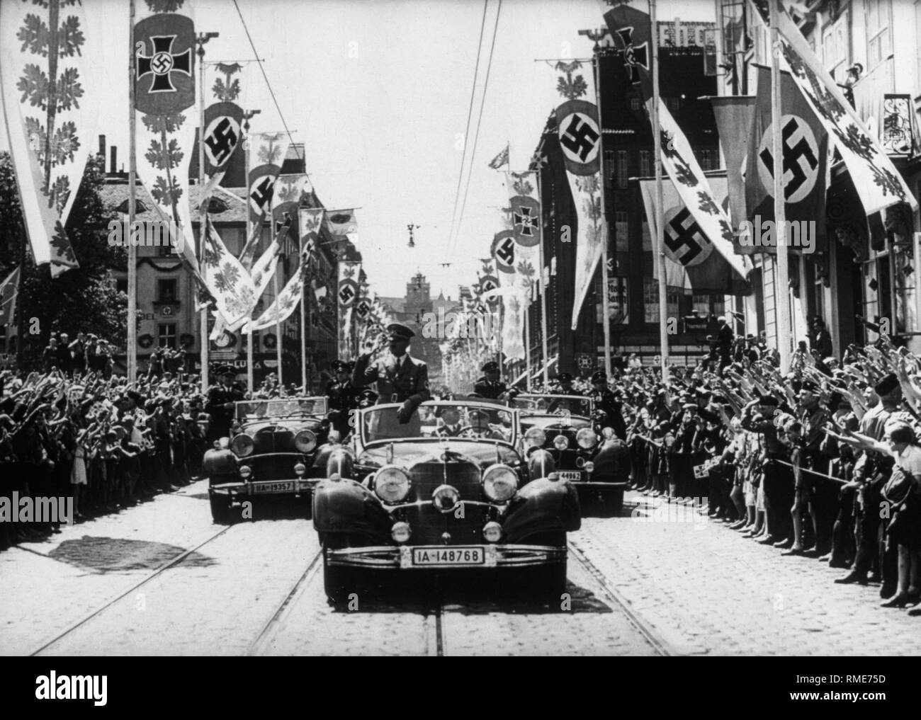 Adolf Hitler in his Mercedes, celebrated by an enthusiastic population. Left and right, street decorated with Nazi flags, including the flag of the National Socialist War Victim's Care next to a swastika flag. Stock Photo