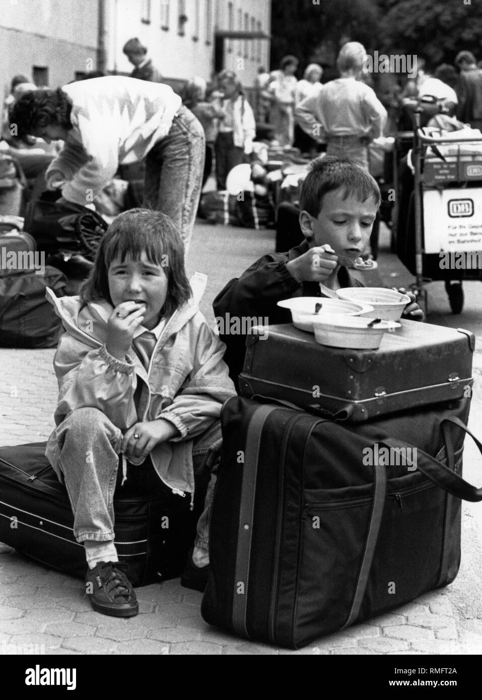 Refugees from the GDR in front of the Federal Reception Center in Giessen, where they are to be registered and receive their admission certificates. In the foreground two children are sitting on suitcases while having a meal. Stock Photo