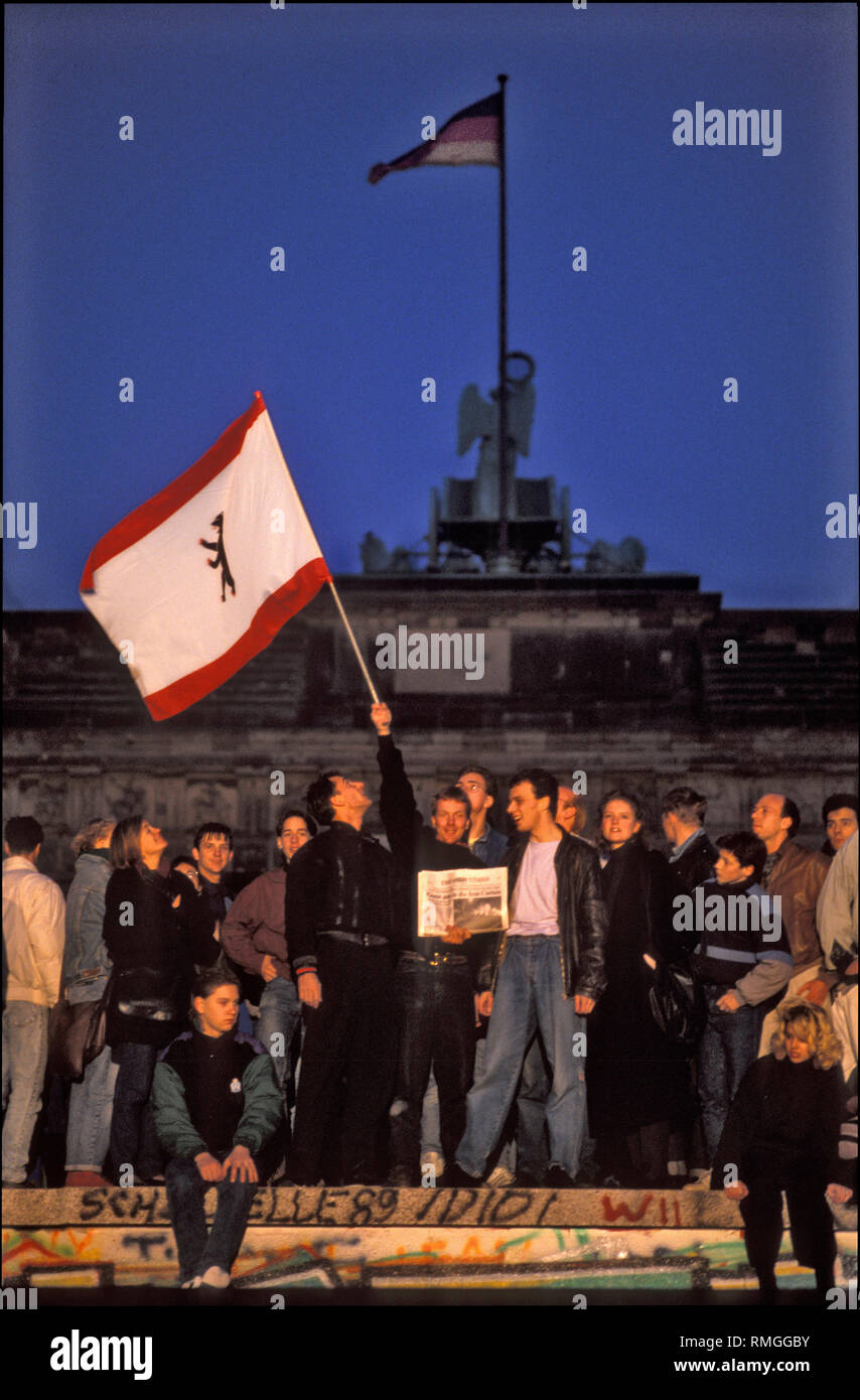 Thousands of Berliners are gathering at the Berlin Wall at the Brandenburg Gate to celebrate the opening of the border. A man is holding up the flag of Berlin. Stock Photo