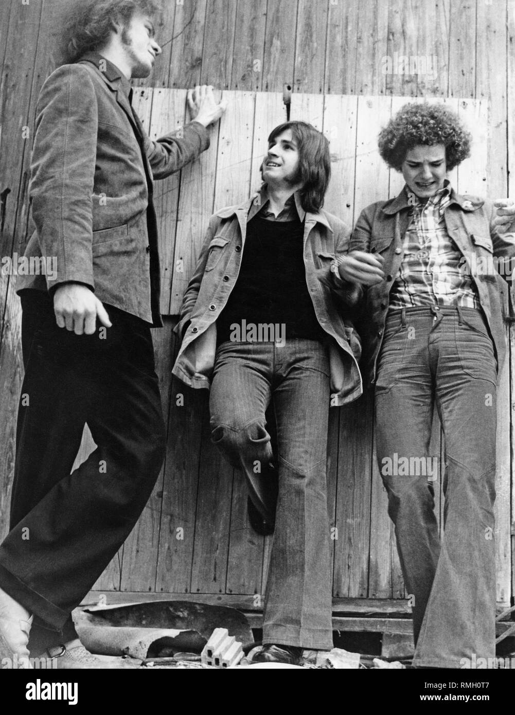 Three teenagers, wearing bell bottoms, lean against a wooden wall. Stock Photo