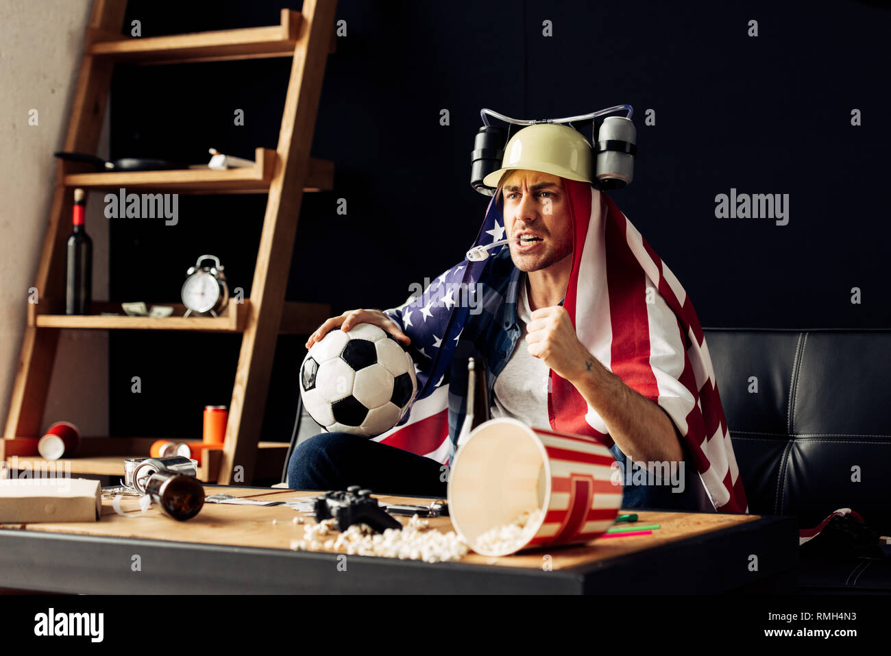 man wearing beer helmet drinking and holding ball while sitting with