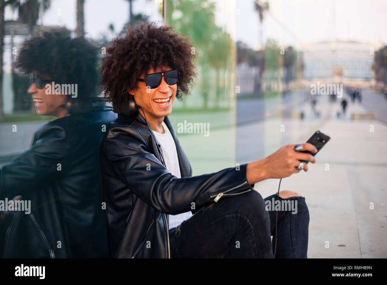 Handsome young black man with afro hairstyle sitting in the street with mobile and skateboard in a sunny city Stock Photo