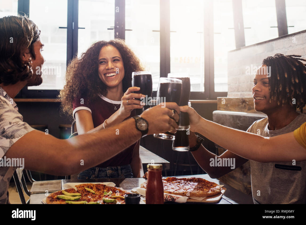Smiling young men and women sitting inside a restaurant toasting soft drinks. Multi-ethnic group of people toasting cold drinks with pizza on table at Stock Photo