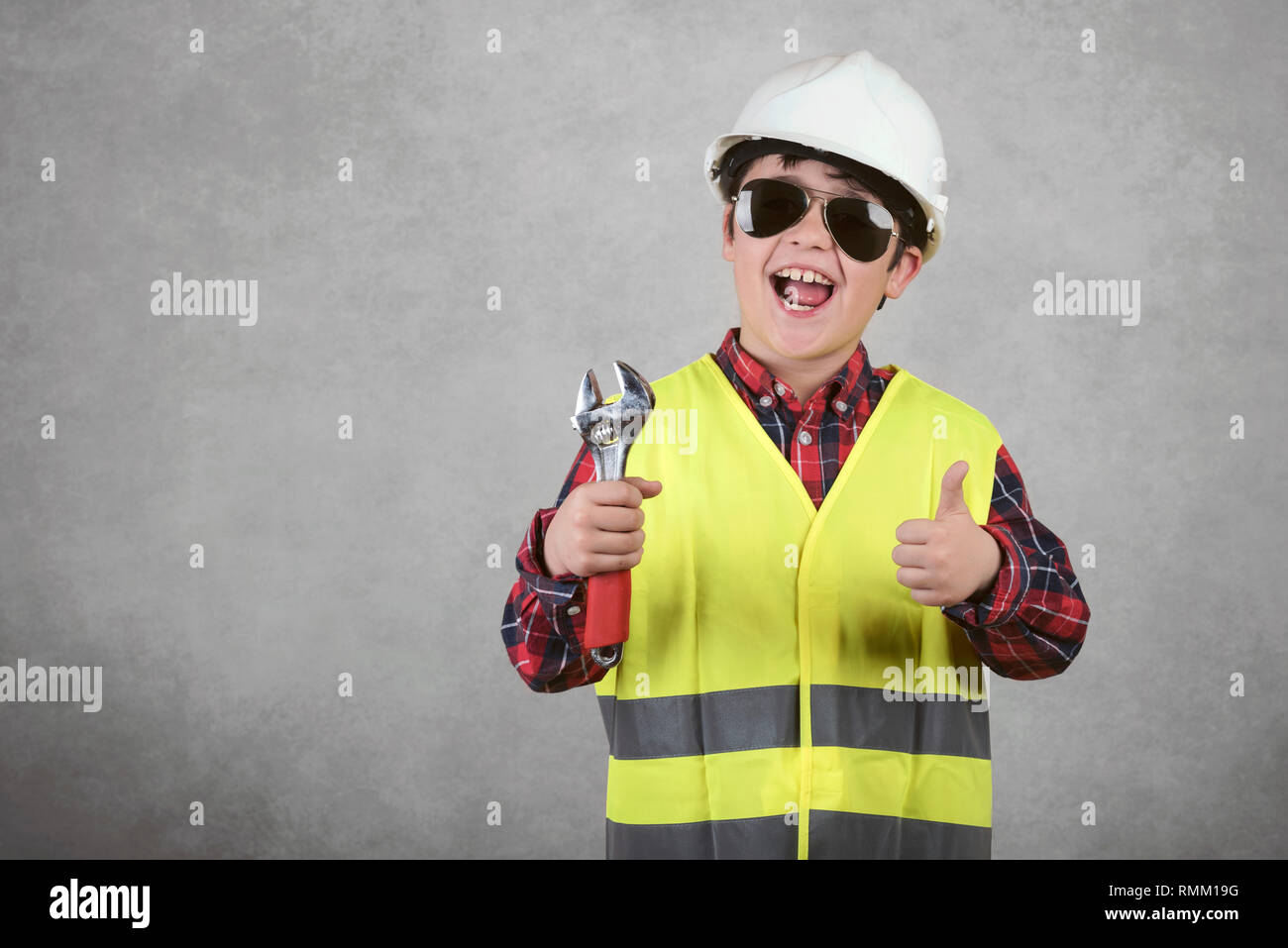 little child construction worker in White helmet and sunglasses and holding a wrench  against gray background Stock Photo