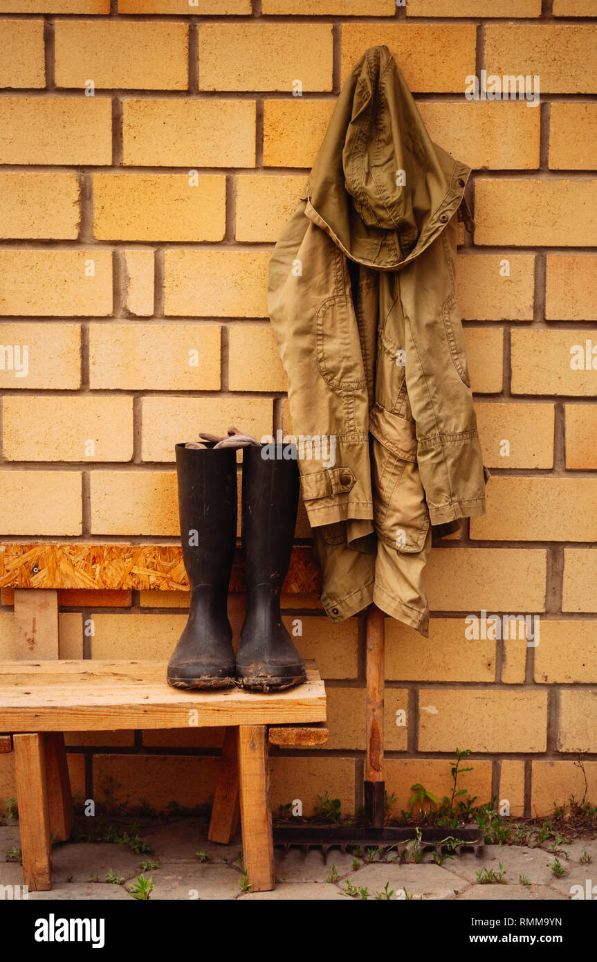 Farmer kit - rain coat haning on rake and rubber boots on a bench. Toned. Stock Photo
