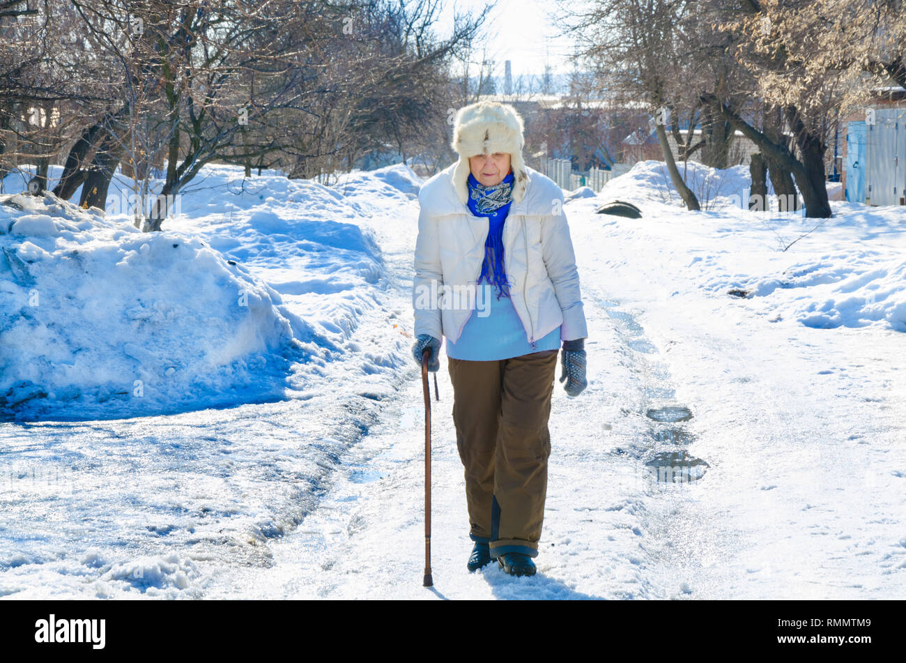 grandmother walk with cane on road. old person leaning on a walking stick. caucasian female senior at cold winter. fashion lady outside. healthy leisure of grandma in fur hat. elderly go on snow. Stock Photo