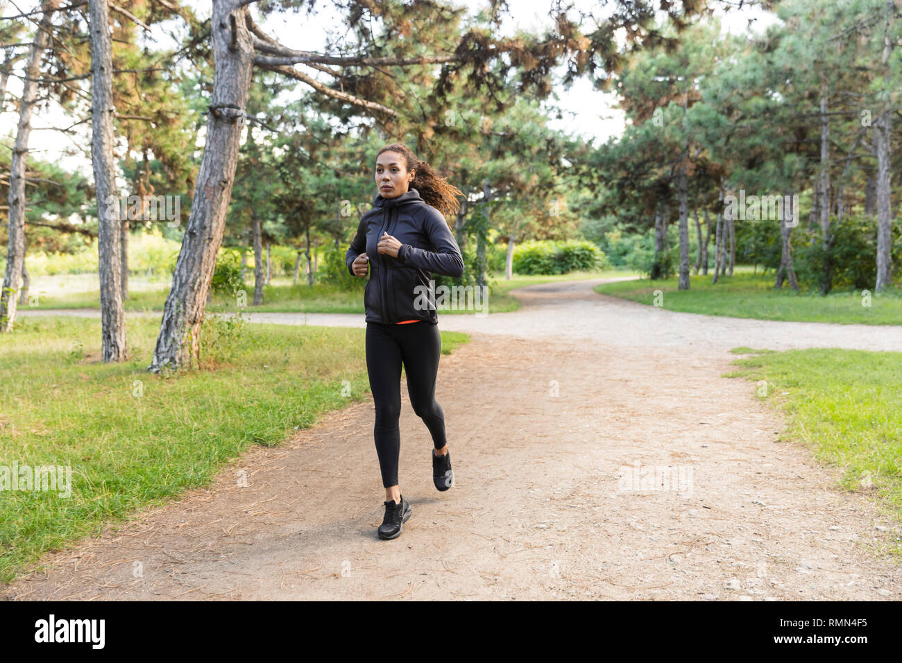 Image of young woman 20s wearing black tracksuit working out while running through green park