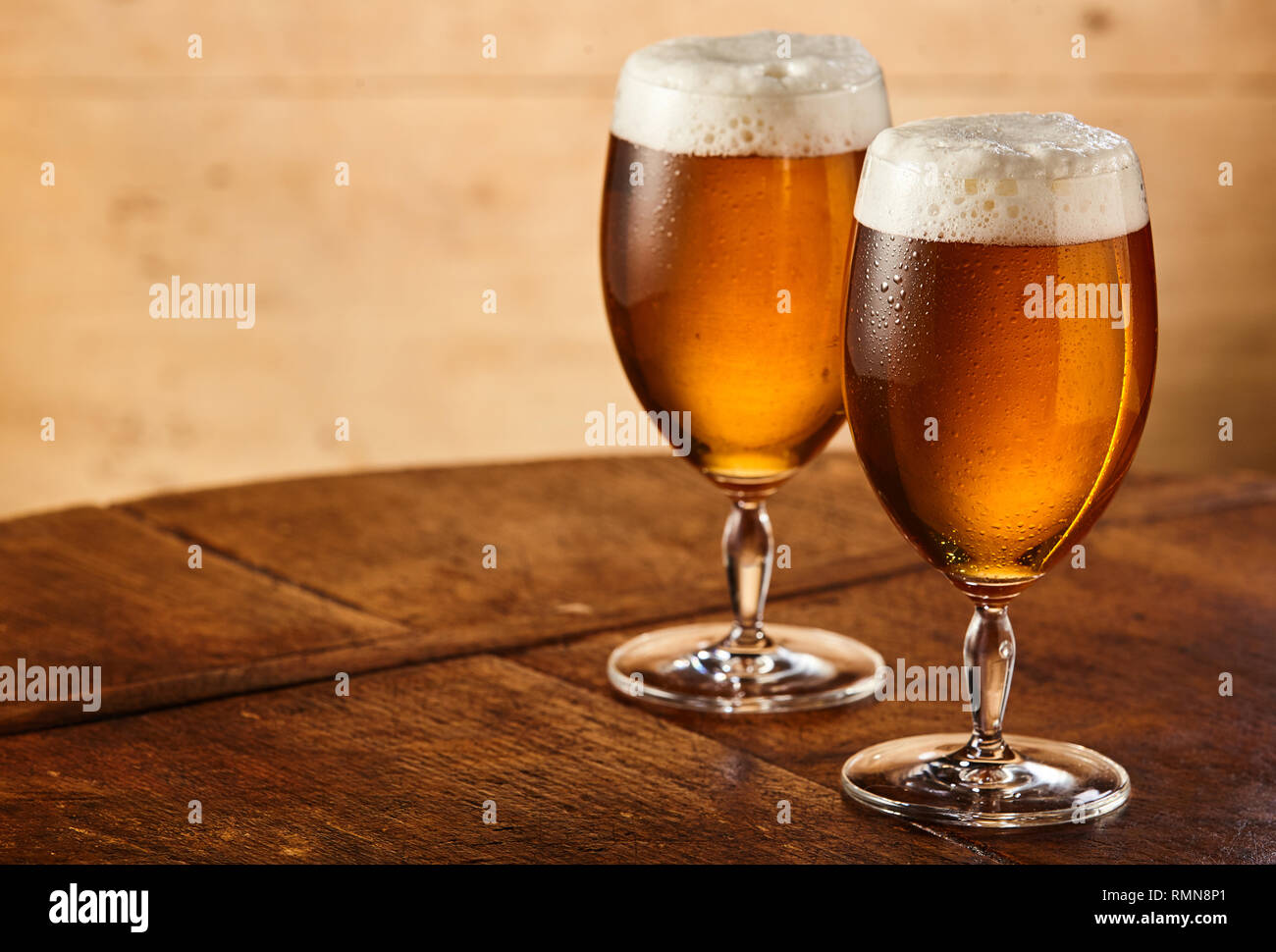 Two tall stemmed glasses of fresh chilled draft or craft beer with fresh frothy heads standing side by side on an old table with lateral copy space Stock Photo