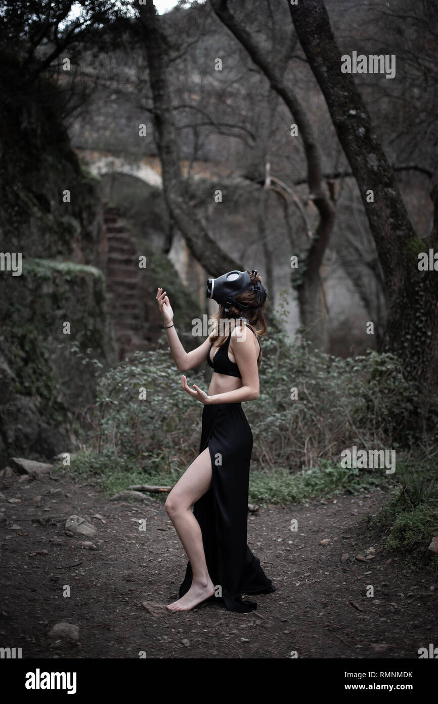 Side view of anonymous barefoot woman in black clothes and gas mask standing in amazing spooky forest Stock Photo