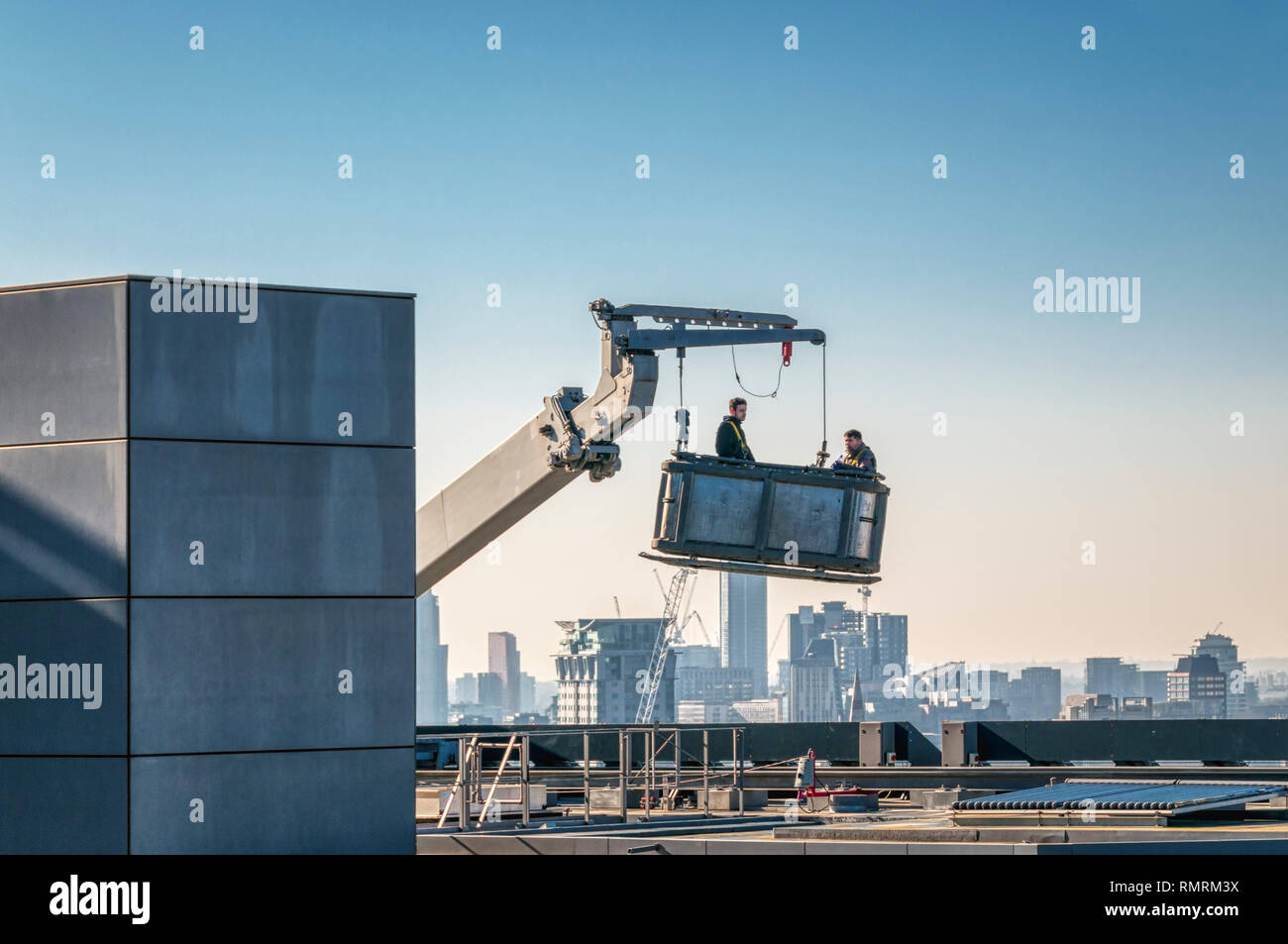 Two workers prepare to descend from the top of a tall building working from an access cradle. City skyline in background. Stock Photo