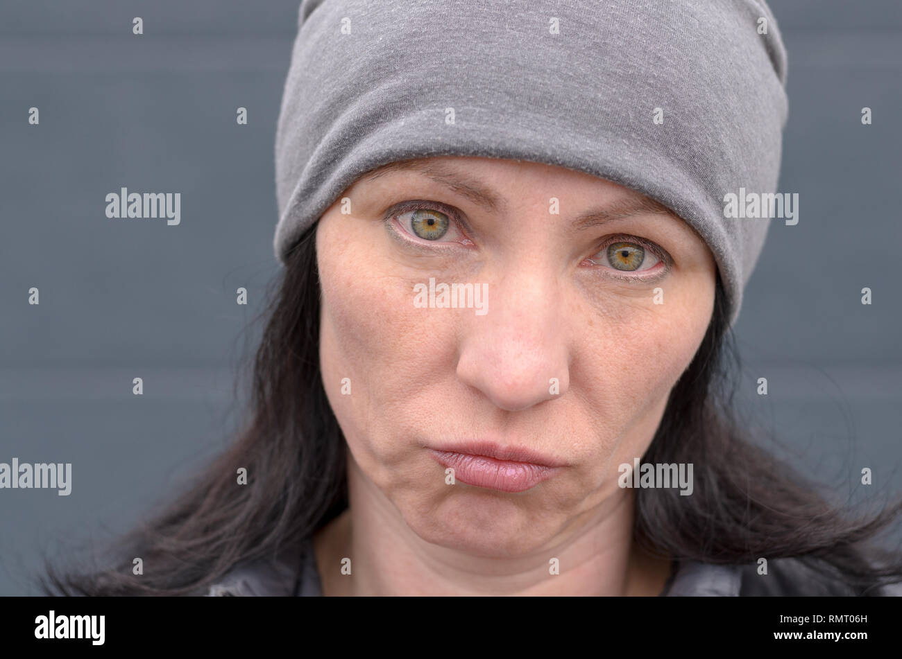 Dubious woman wearing a knitted grey winter cap grimacing and looking at camera with a sad expression in a close up cropped portrait Stock Photo