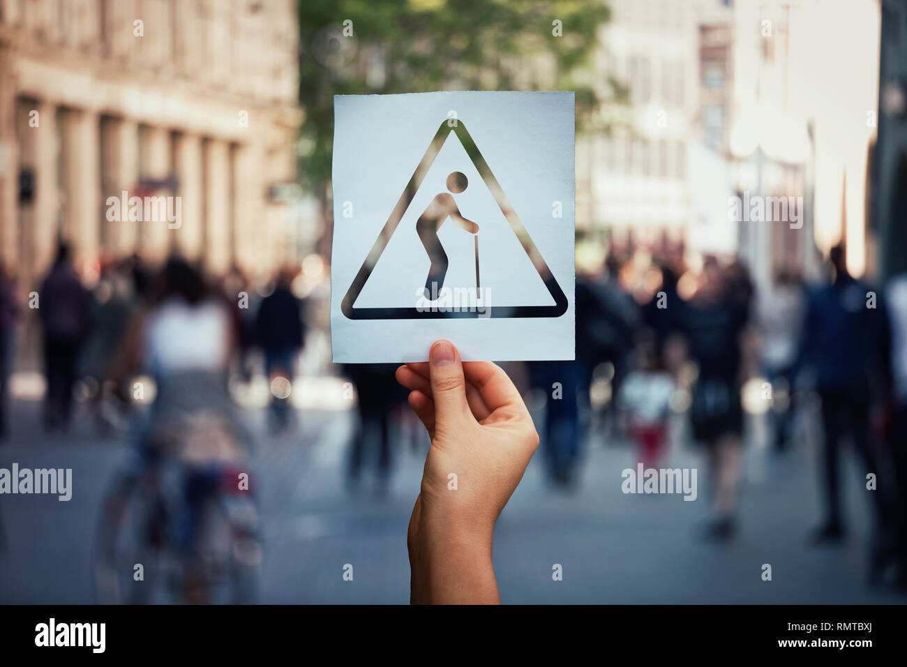 Global population ageing problem. Hand holding a paper sheet as an old man using walking stick over a crowded street background. World growth of the n Stock Photo