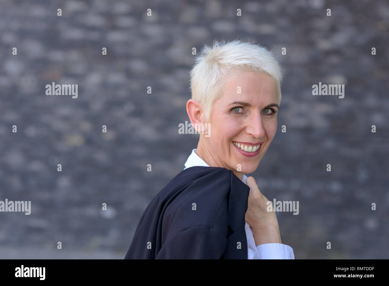 Happy woman is wearing her jacket over her shoulder in front of a black wall Stock Photo