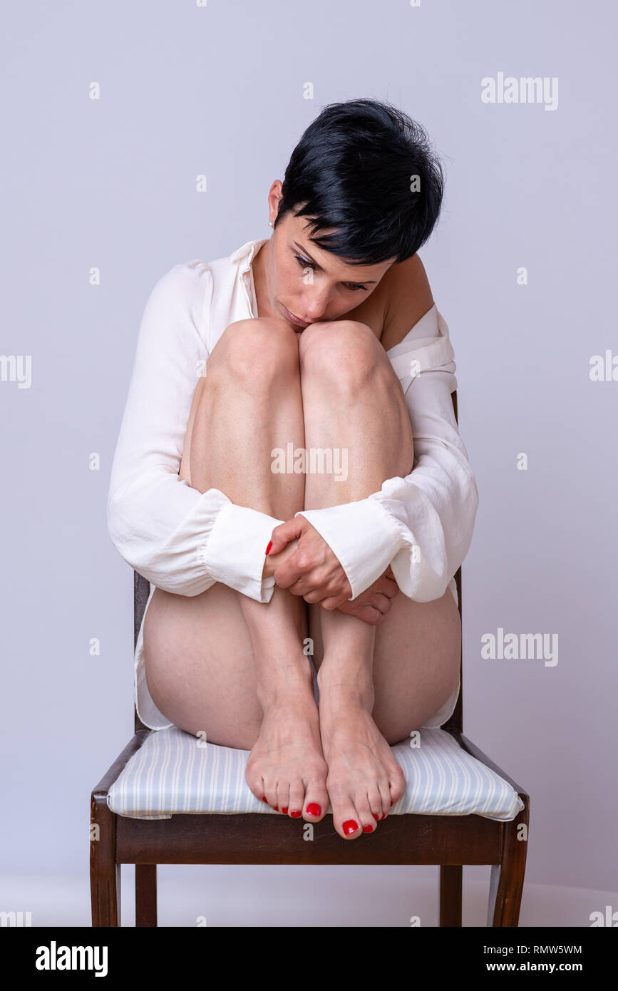 Lonely depressed woman sitting on a chair resting her chin on her bent knees as she stares disconsolately down at the floor Stock Photo