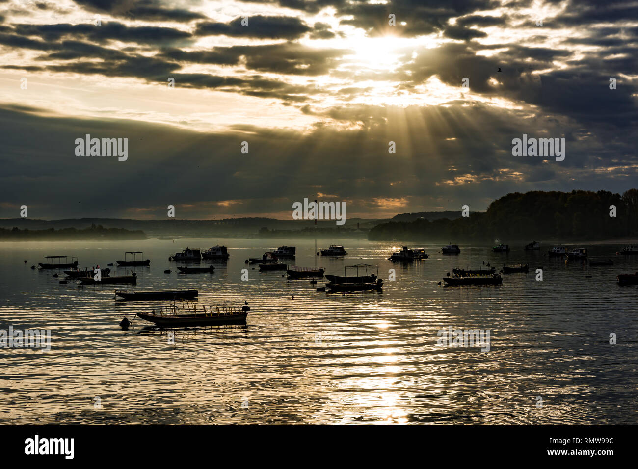 The morning sun's rays that pierce through the clouds, illuminate the boats parked on Zemun quay on river Danube. Stock Photo