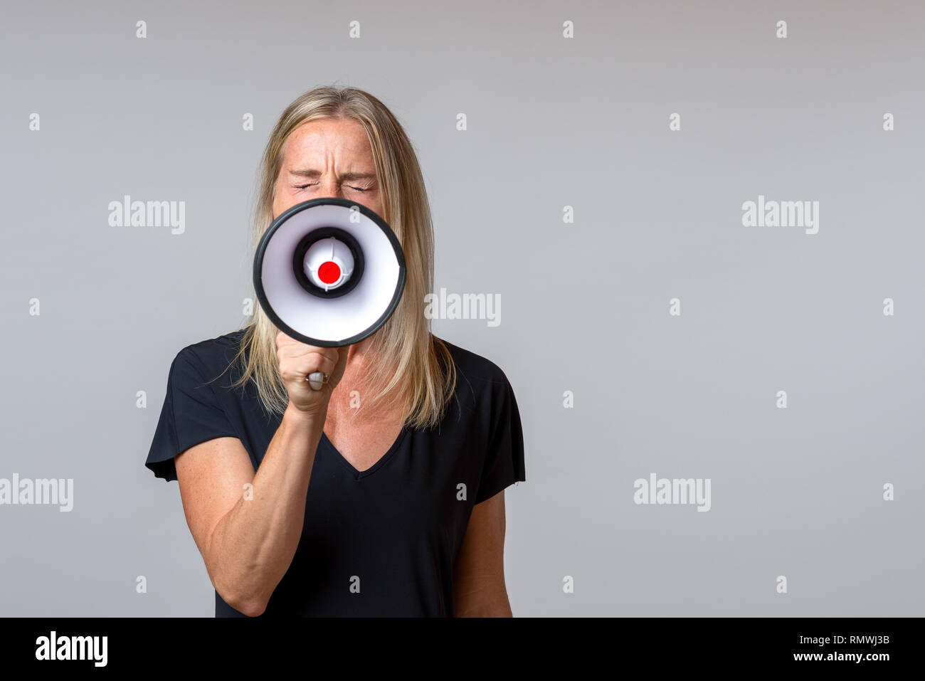 Angry woman yelling into a handheld megaphone in a concept of activism, strike, demonstration, protest or public speaking over grey with copy space Stock Photo