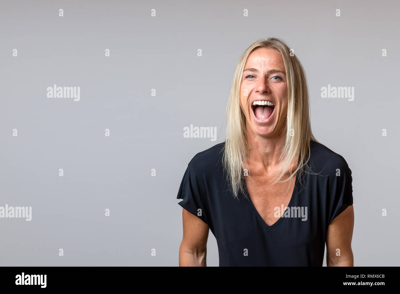 Happy temperamental mature blond woman yelling and glaring at the camera with her mouth wide open isolated on grey with copy space Stock Photo