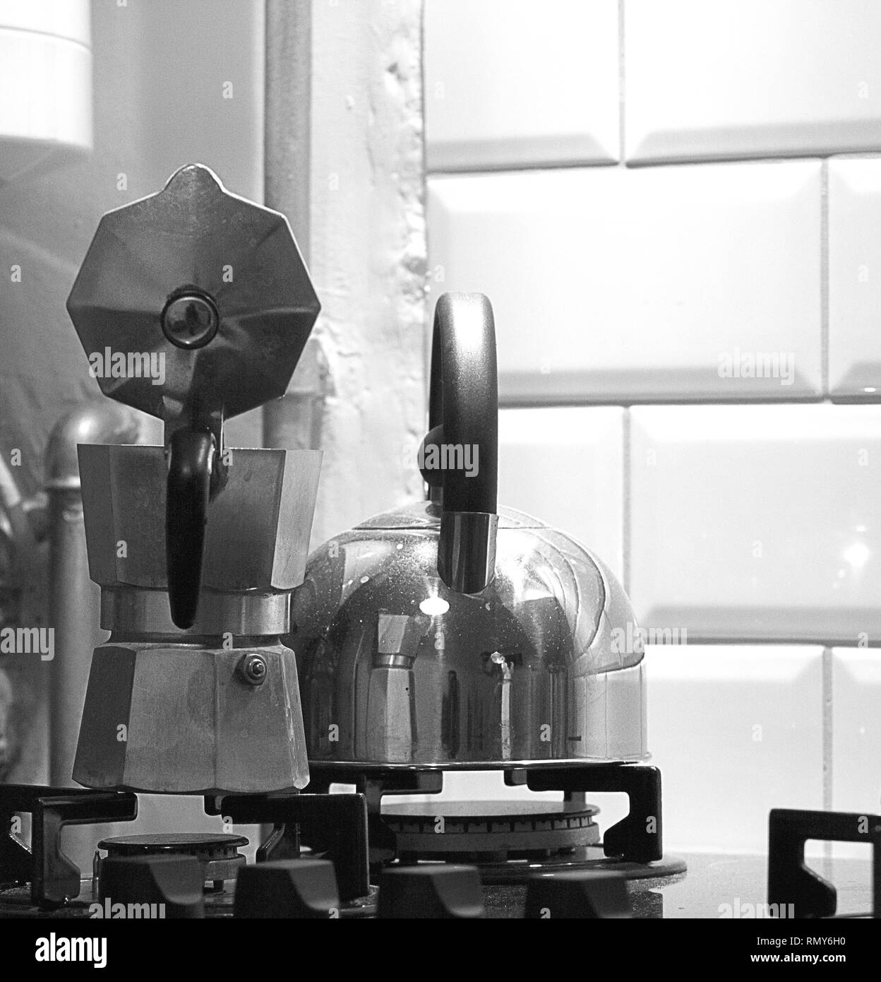 Black and white close-up of an aluminum moka pot and a steel kettle with bakelite handles on a gas stove with white retro ceramic wall tiles. Stock Photo