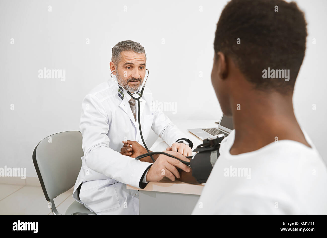 Doctor using sphygmomanometer for measuring blood pressure of patient. African man sitting on consultation, holding hand on table. Doctor wearing in medical white gown. Stock Photo