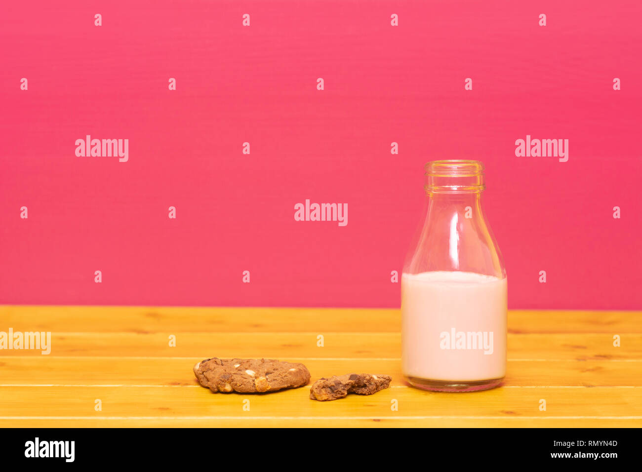 One-third pint glass milk bottle half full with strawberry milkshake and a half-eaten chocolate chip cookie, on a wooden table against a pink backgrou Stock Photo