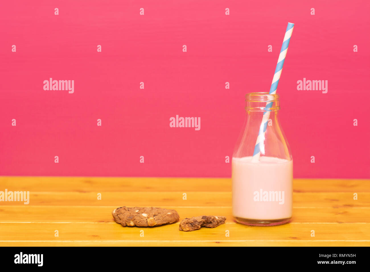 One-third pint glass milk bottle half full with strawberry milkshake with a retro straw and a half-eaten chocolate chip cookie, on a wooden table agai Stock Photo