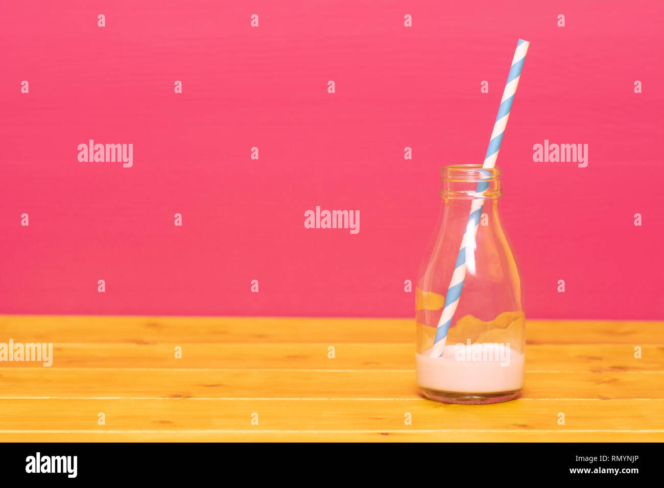 One-third pint glass milk bottle with dregs of strawberry milkshake and a retro paper straw, on a wooden table against a pink background Stock Photo