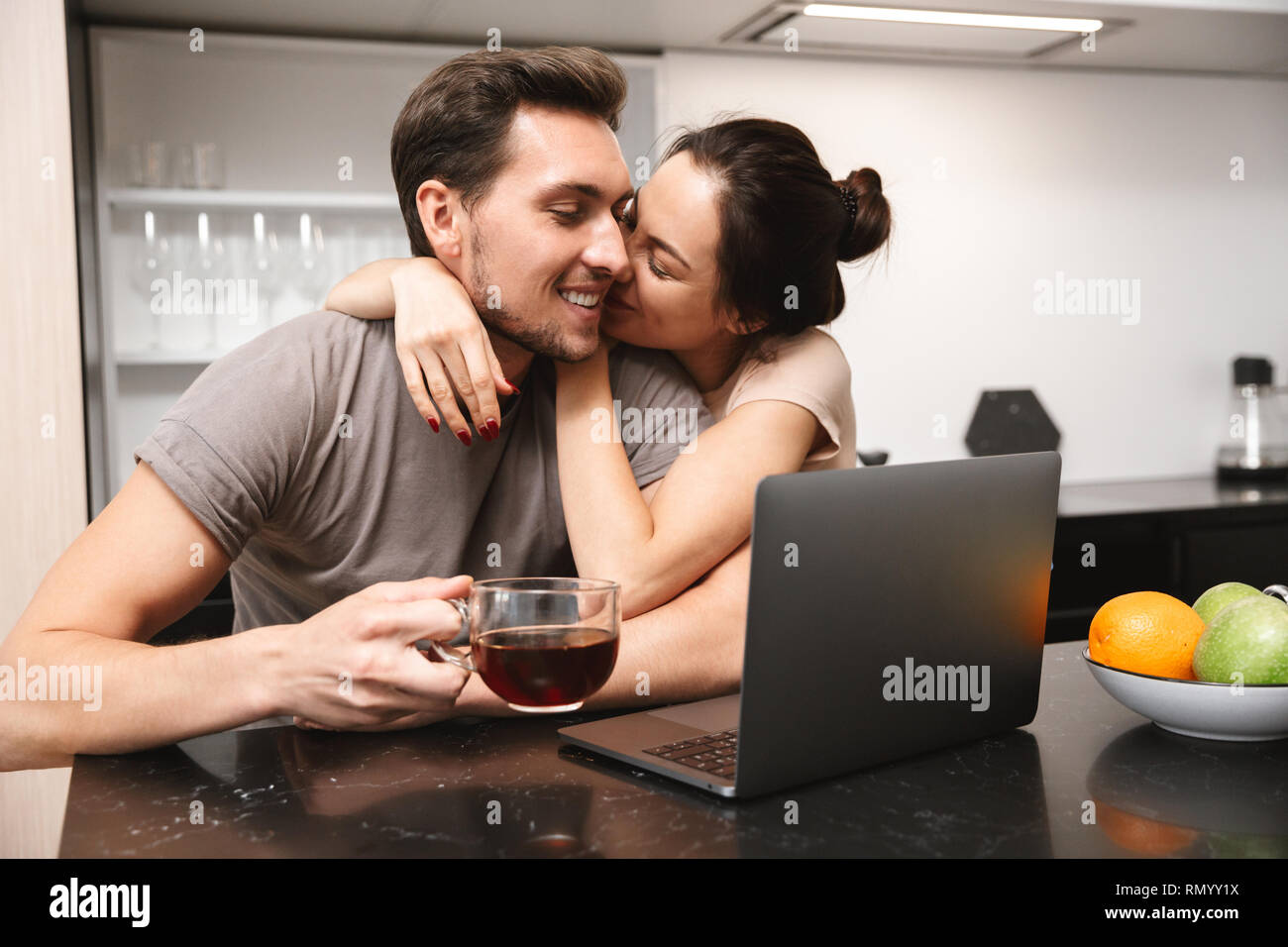 Photo of jouful couple man and woman sitting in kitchen and using laptop while having breakfast together Stock Photo