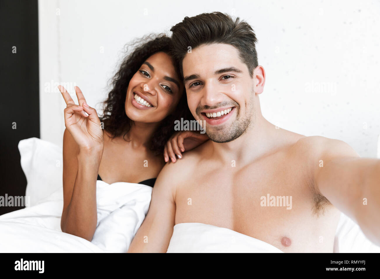 Beautiful happy young multiethnic couple relaxing in bed under blanket, taking a selfie Stock Photo