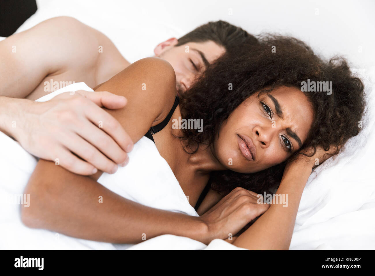 Beautiful young multiethnic couple relaxing in bed under blanket, woman frowning Stock Photo