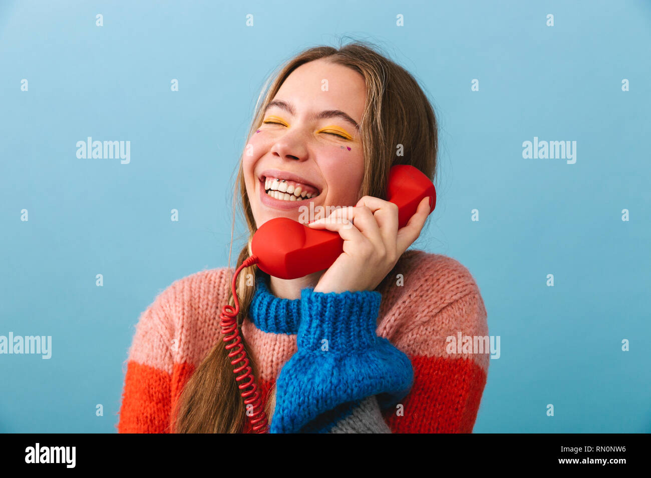 Excited young girl wearing winter clothes standing isolated over blue background, talking on a landline phone Stock Photo