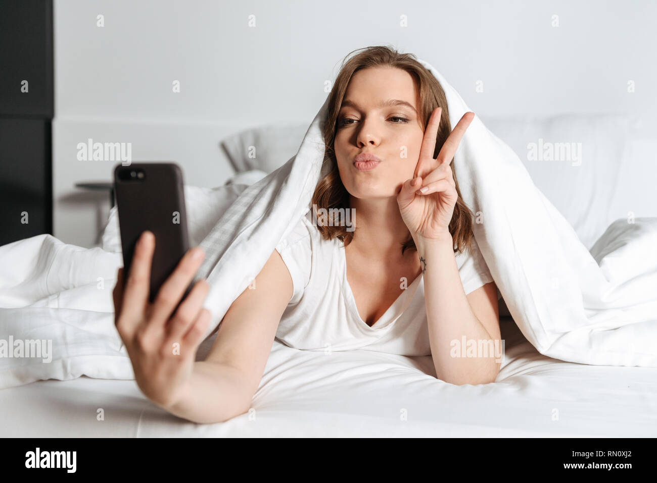 Attractive smiling young woman lying in bed under the blanket at home, taking a selfie Stock