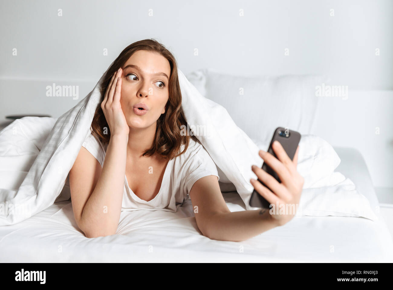 Attractive smiling young woman lying in bed under the blanket at home, taking a selfie Stock