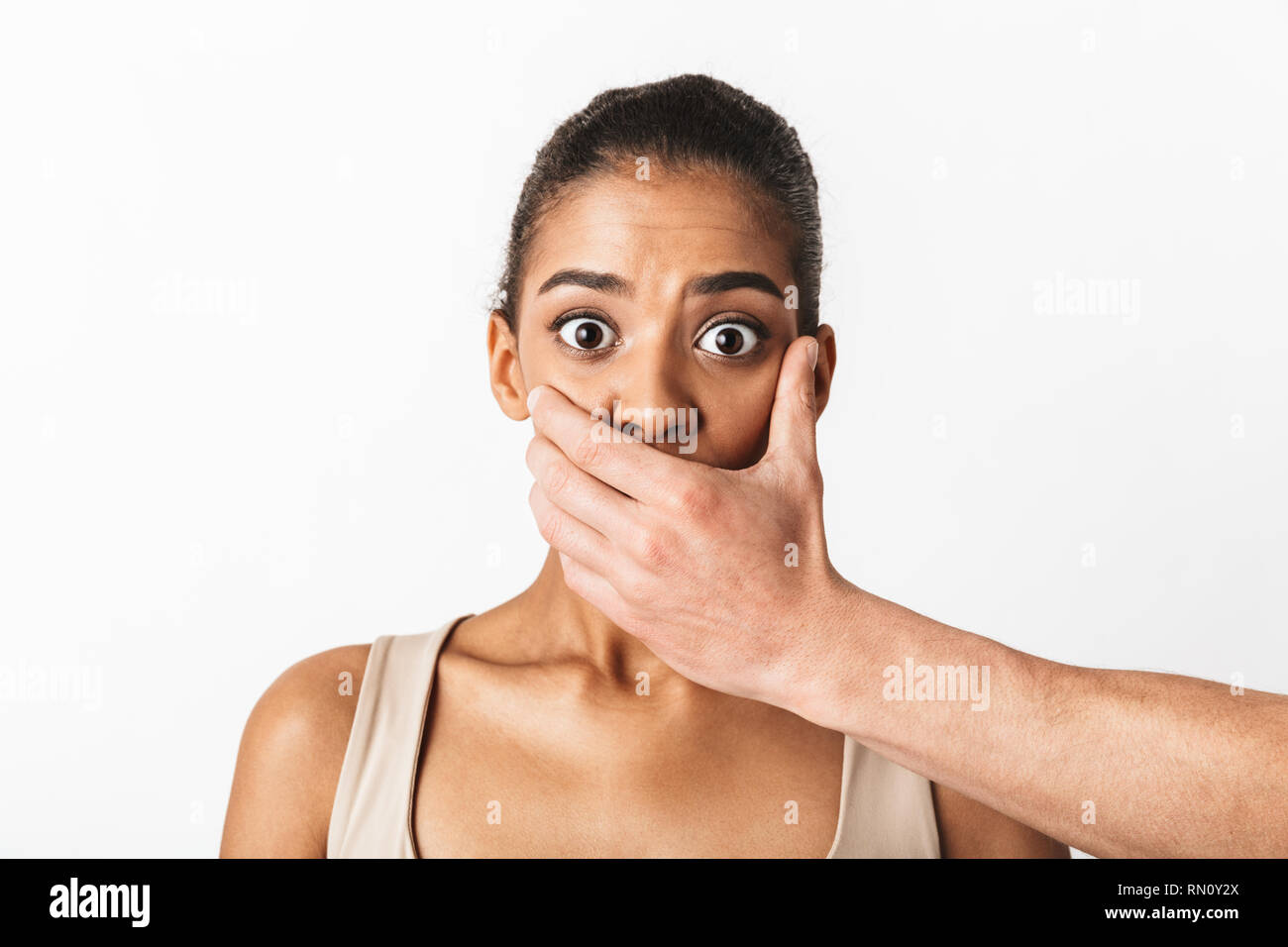 Scared young african woman looking camera while man's hand covering her mouth isolated over white background Stock Photo