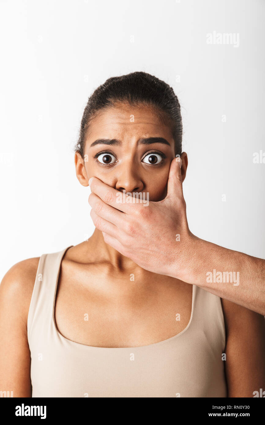 Scared young african woman looking camera while man's hand covering her mouth isolated over white background Stock Photo