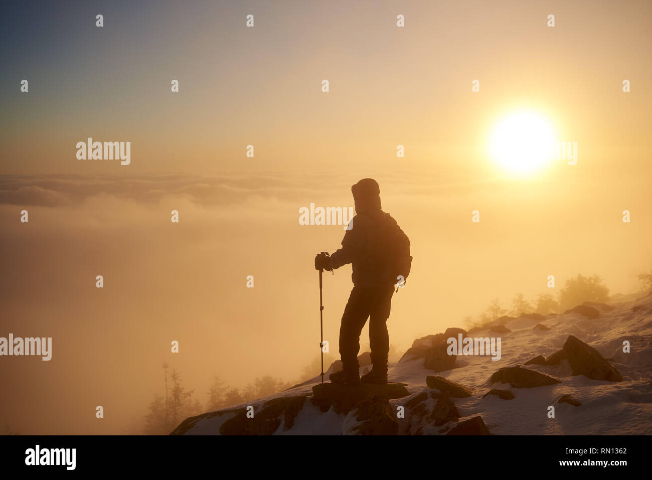 Back view of tourist hiker with backpack and hiking sticks resting on rocky mountain peak on copy space background of beautiful foggy valley filled with white puffy clouds at dawn at winter. Stock Photo