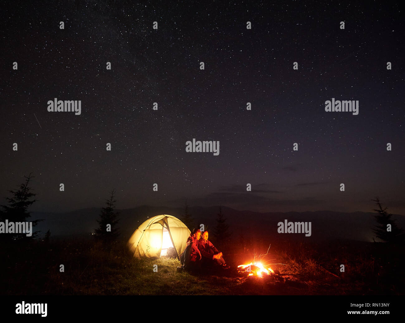 Romantic Couple Tourists Boy And Girl Sitting Near Illuminated Tent By Burning Bonfire Under Starry Sky Enjoying Quiet Night Camping In Mountains Tourism Traveling And Beauty Of Nature Concept Stock Photo