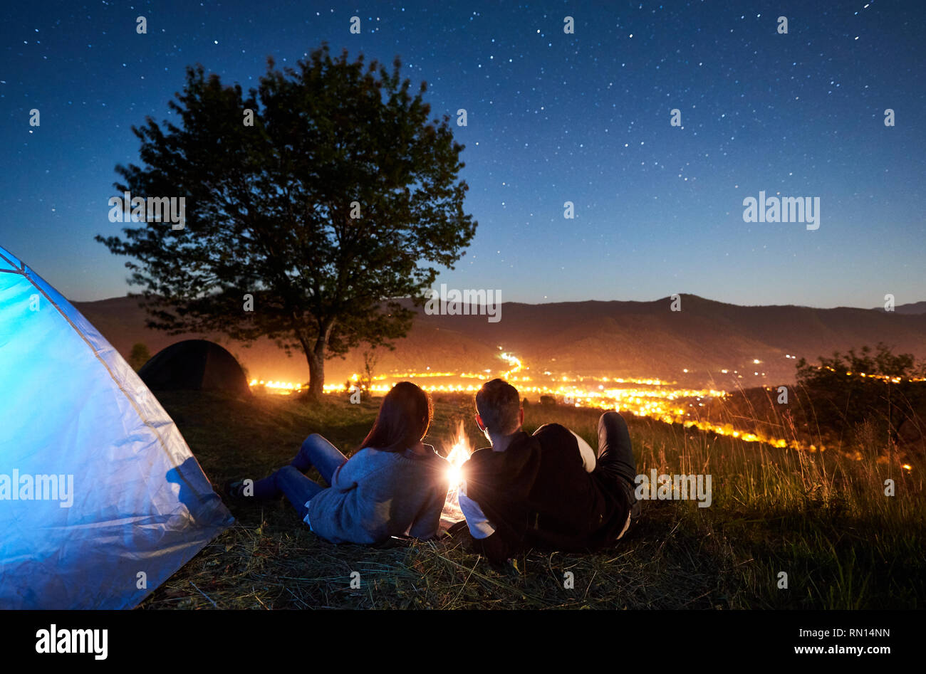 Rear view of happy romantic couple travellers having a rest at bonfire near illuminated blue tourist tent under night sky full of stars. On background starry sky, mountains, big tree and luminous city Stock Photo