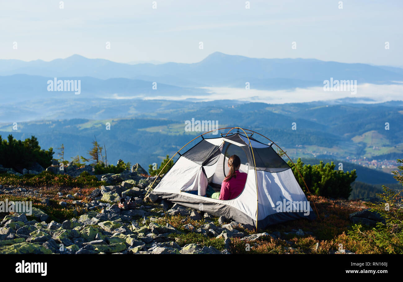 Camping on the top of mountain on bright summer morning. Back view of woman sitting in the entrance of tourist tent. On foggy mountains background. Tourism adventure holiday active lifestyle concept Stock Photo