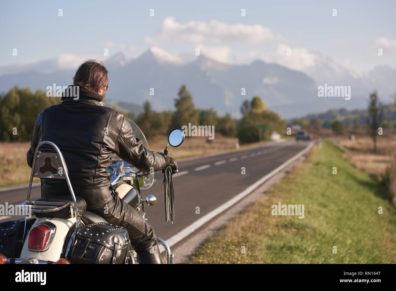 Back view of biker in black leather jacket riding cruiser motorcycle along road on blurred background of beautiful mountain range with snowy peaks and blue sky on bright sunny autumn day. Stock Photo
