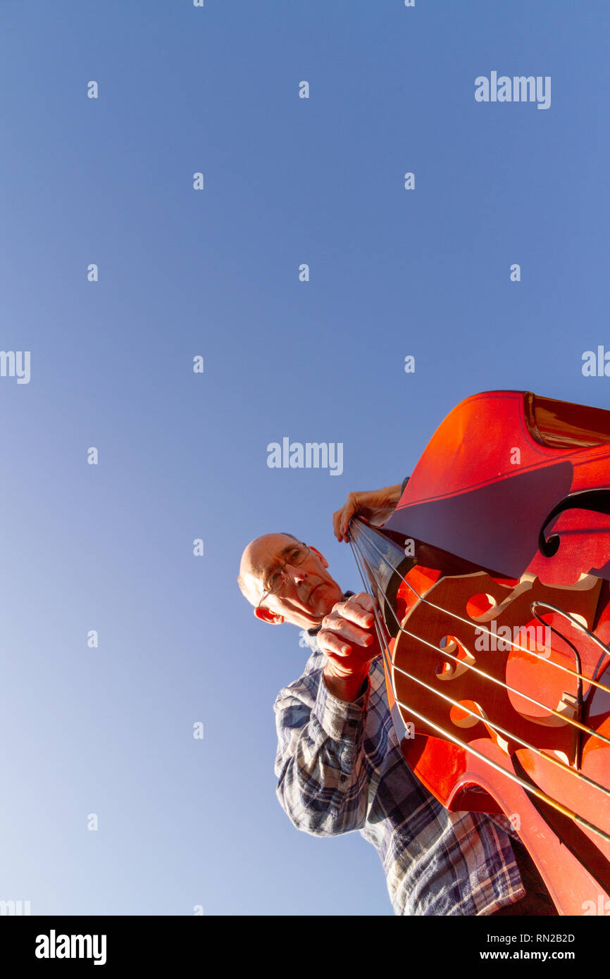 Older male person playing double bass, shot from below against a blue sky (with space for copy). Portrait orientation Stock Photo