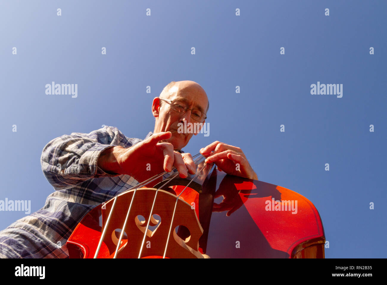 Older male person playing double bass, shot from below against a blue sky (with space for copy). Landscape orientation Stock Photo