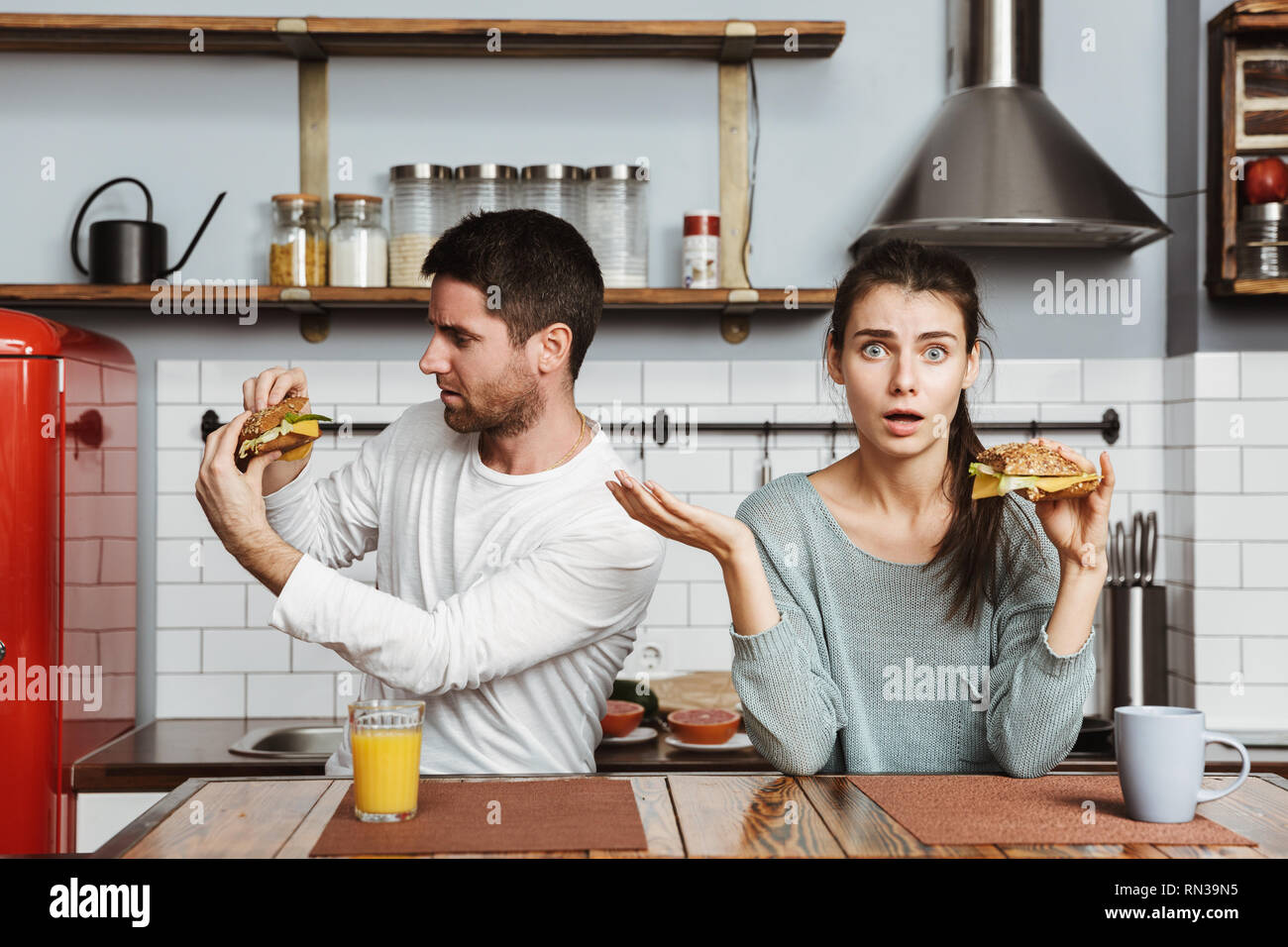 Disgusted young couple sitting at the kitchen during breakfast at home, holding sandwiches Stock Photo