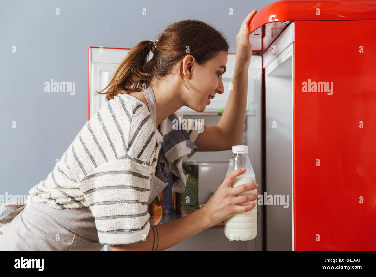 https://c8.alamy.com/comp/RN3AAH/happy-young-woman-standing-at-the-kitchen-at-home-taking-bottle-of-milk-from-a-fridge-RN3AAH.jpg