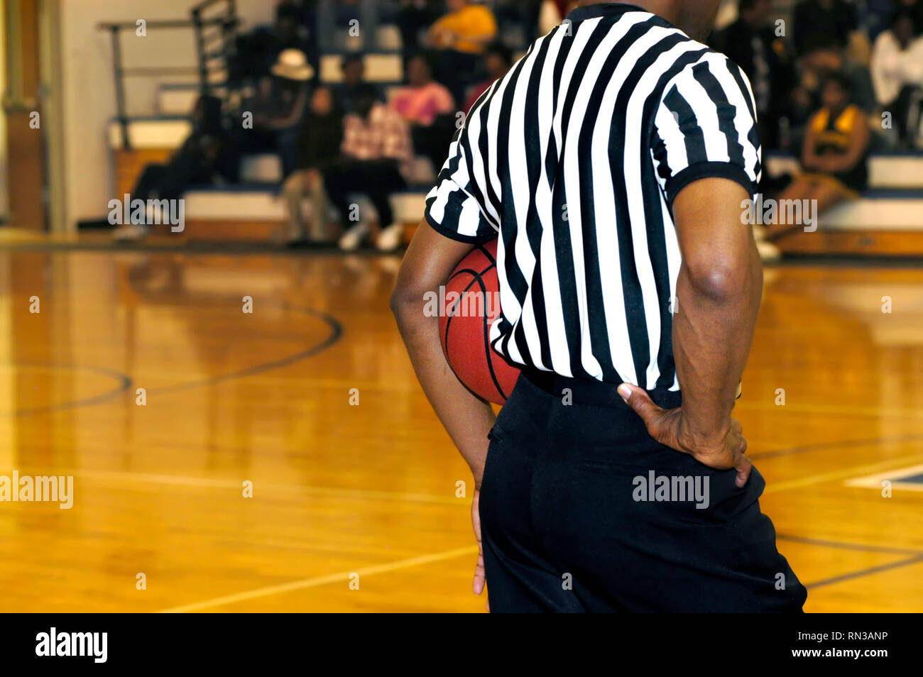 Referee stands mid court holding basketball under his arm. He is wearing the usual black and