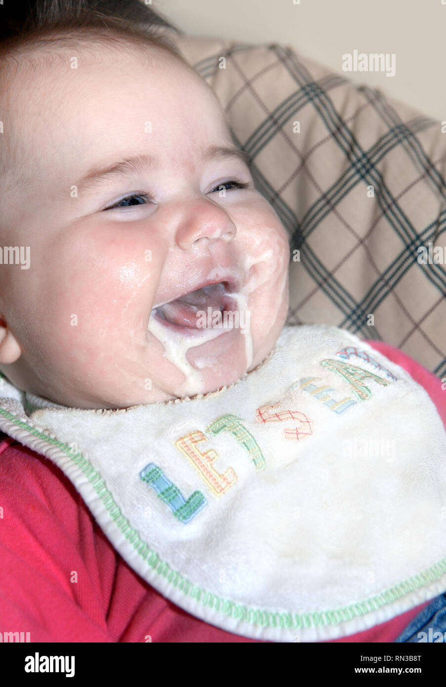 Baby girl, despite having taken a bite of her baby food, grins with every ounce of her being.  Mess ensues with liquid food everywhere. Stock Photo