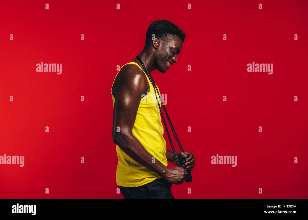 Side view of african muscular man with jumping rope around his neck on red background. Smiling male fitness model with skipping rope. Stock Photo